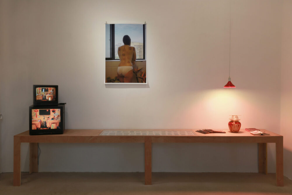 Instalación de Karean Paulina Biswell en porcelana. Sobre la mesa pequeñas polaroids, un jarrón libros y televisión. En la pared un retrato de mujer mostrando su espalda