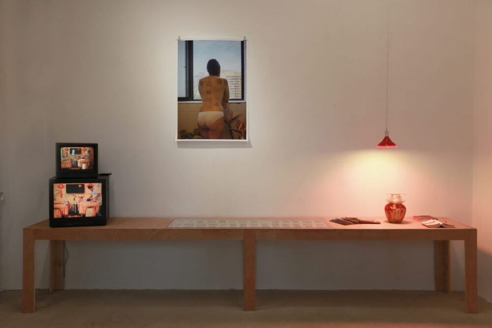 Karean Paulina Biswell installation in china. On the table small polaroids, a vase books and television. On the wall a woman portrait showing her back