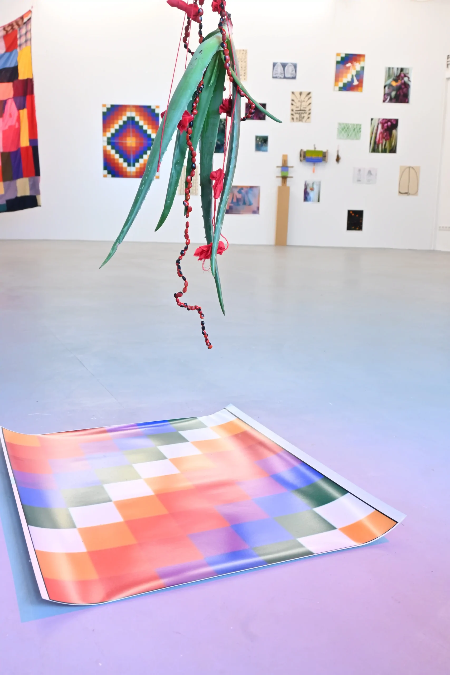 KIMA exhibition view with a suspended aloe plant and beads above a colorful geometric floor work