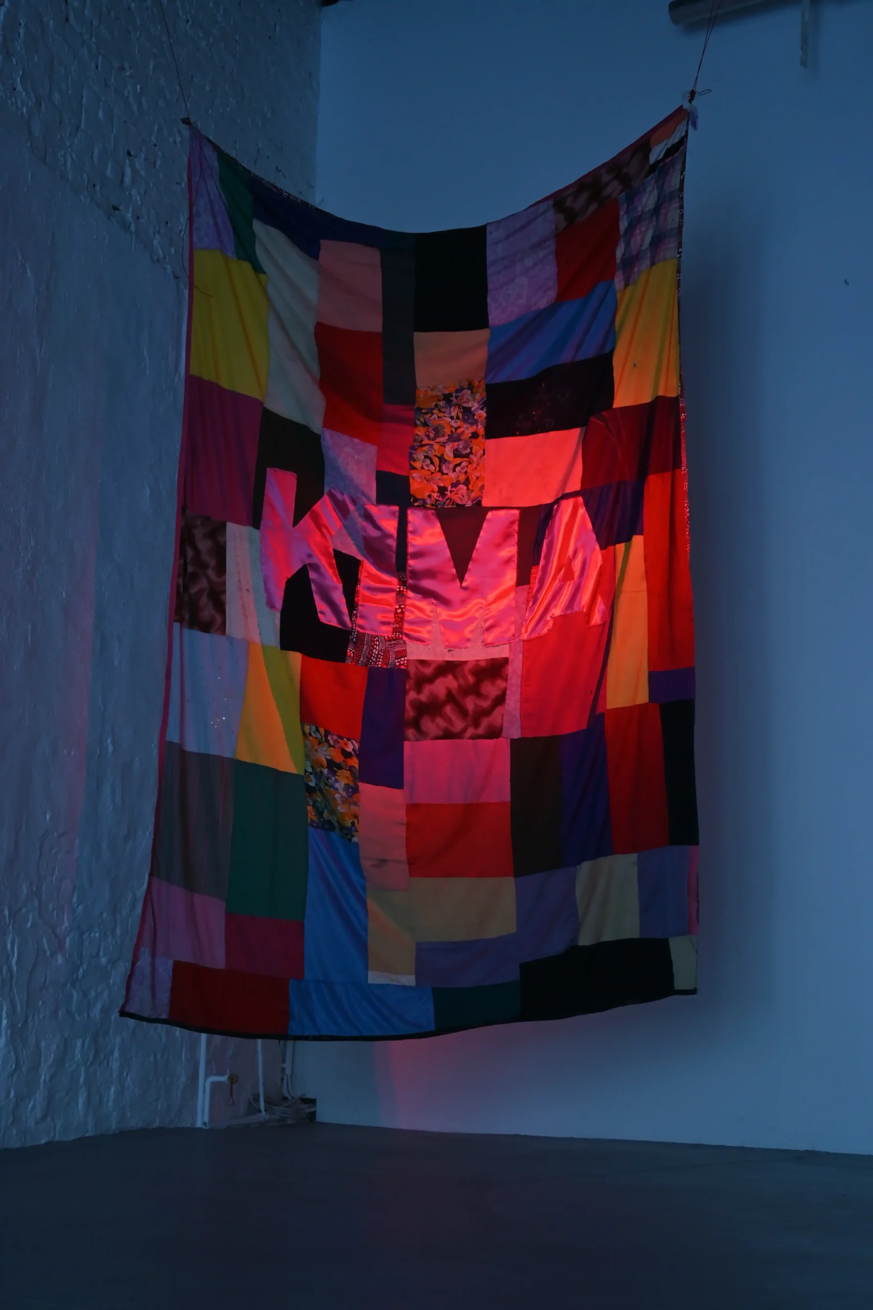 KIMA exhibition view of a suspended multicolored patchwork textile illuminated in red and blue