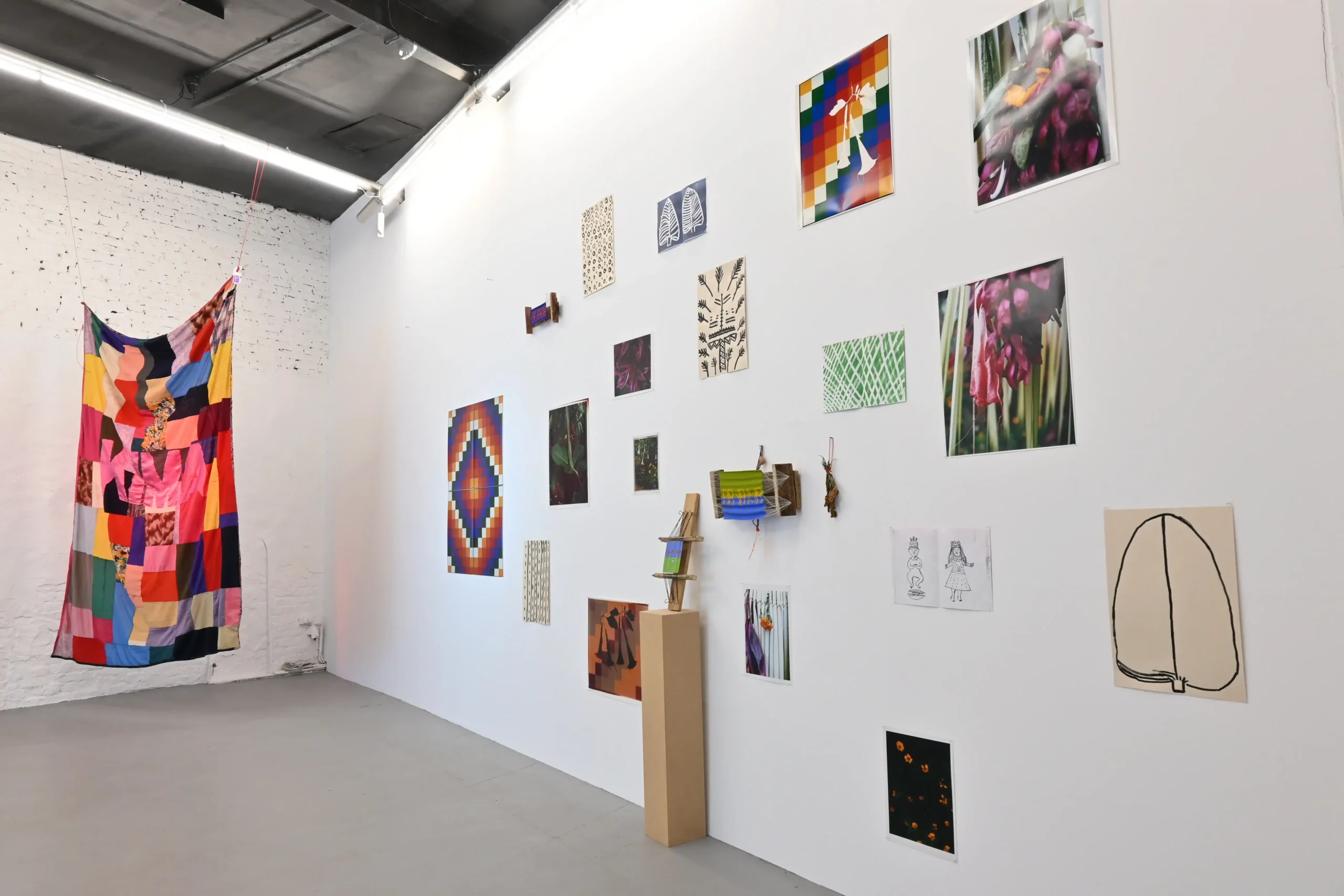 Wide KIMA exhibition view with a suspended patchwork textile and a salon-style wall of artworks