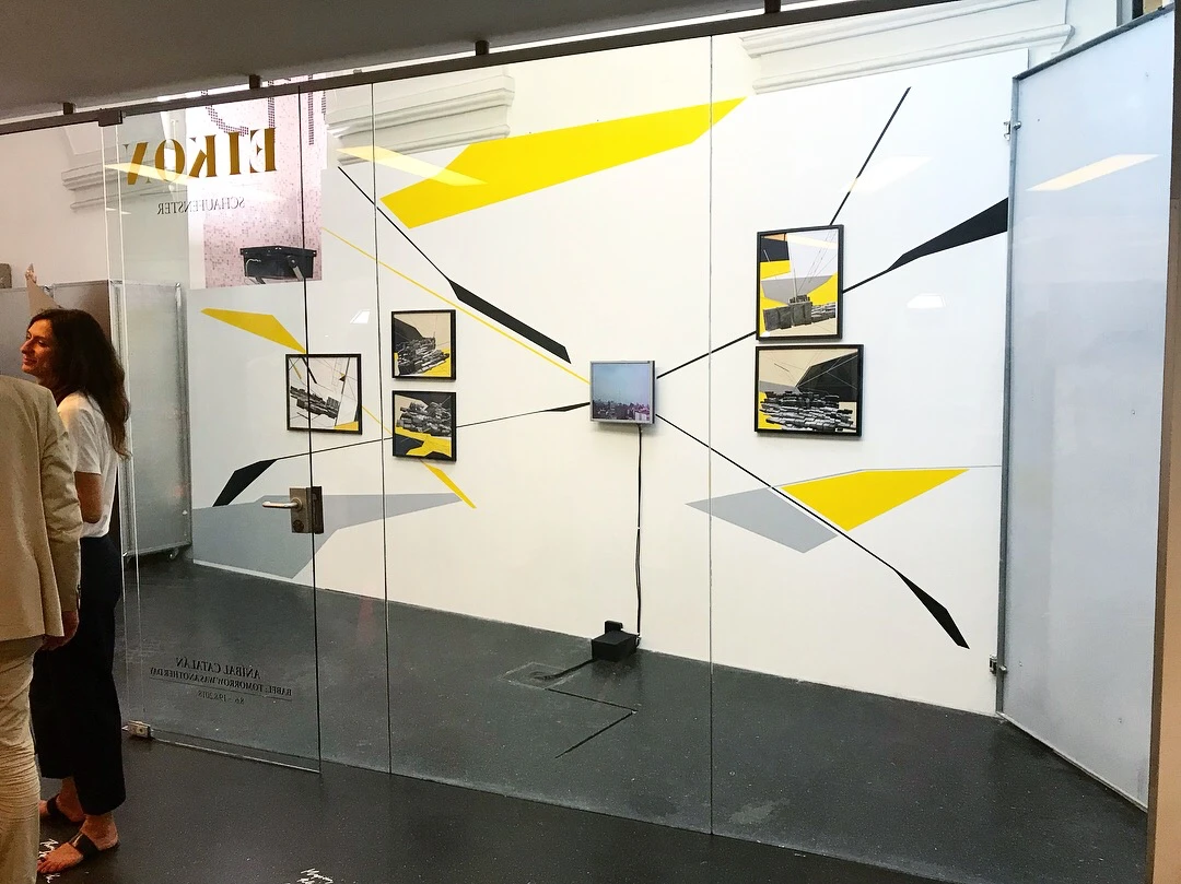 La instalación de Anibal Catalan, Babel: Tomorrow Was Another Day, vista a través del escaparate del EIKON Schaufenster en Viena: una pintura mural en amarillo, negro y gris con collages enmarcados y un pequeño monitor.