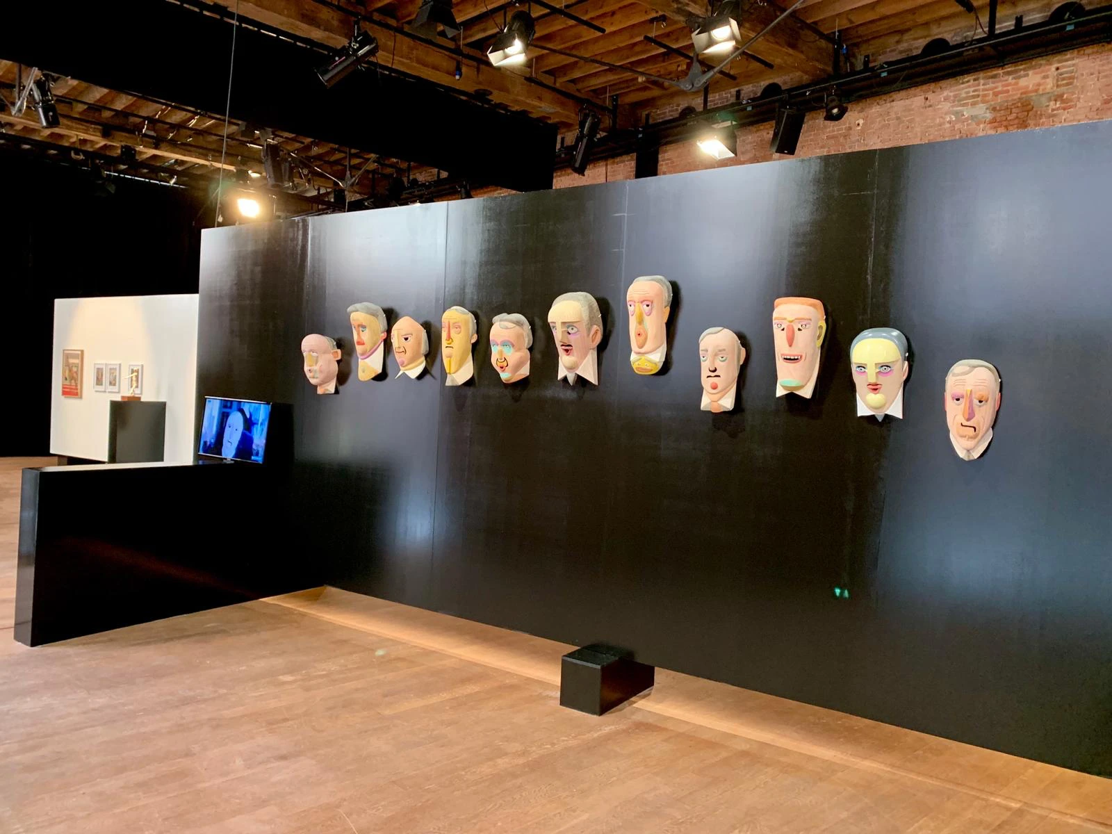 Vista de instalación de las máscaras de madera talladas y pintadas de Mauricio Limón sobre una pared negra, con una pantalla de video y el mostrador de wildpalms, POPPOSITIONS Art Fair, Bruselas