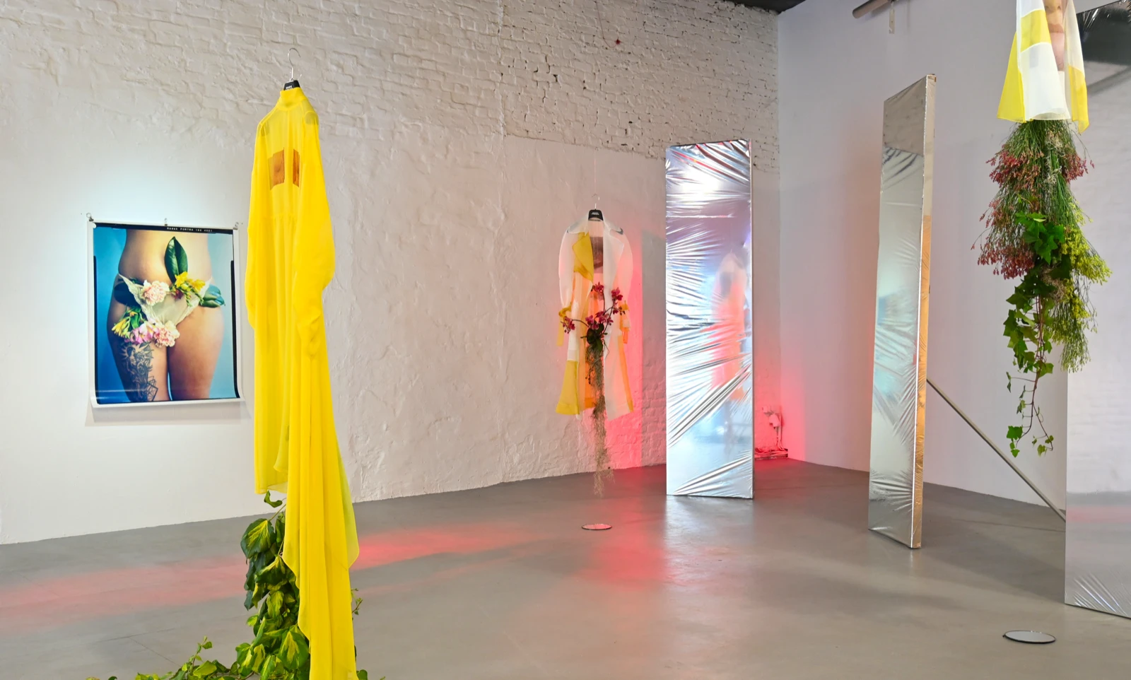 Installation view with a yellow sheer robe entwined in ivy hanging from a wall hook, a framed photograph of a tattooed thigh dressed with fresh flowers, two mirrored panels, and a yellow-and-white fabric piece hanging from the ceiling, lit in red.