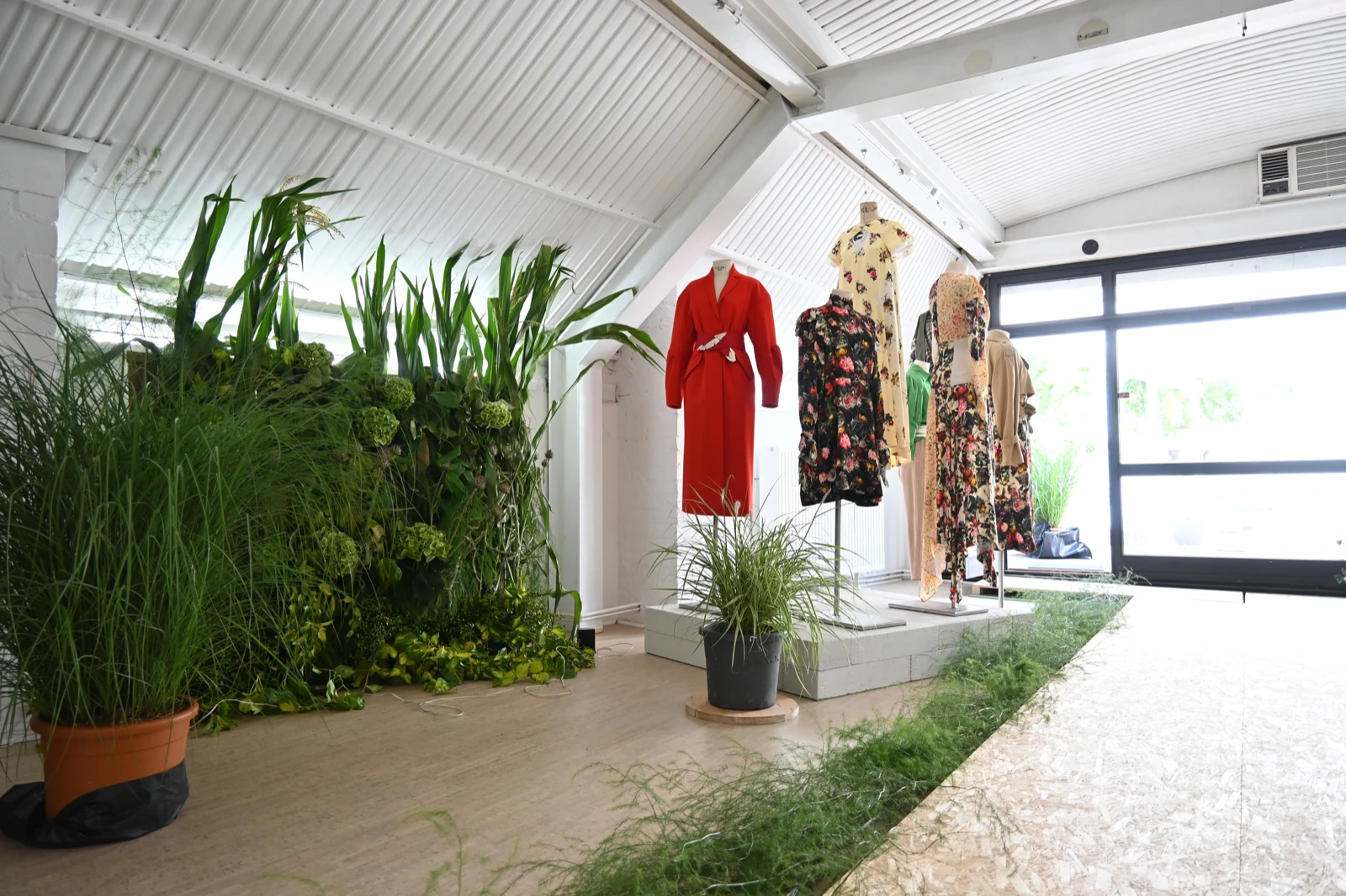 Installation view of Société Angelique’s A Utopian Dream at wildpalms, showing a living wall of plants beside garments — a red coat and floral dresses — displayed on mannequins and hangers near the gallery entrance