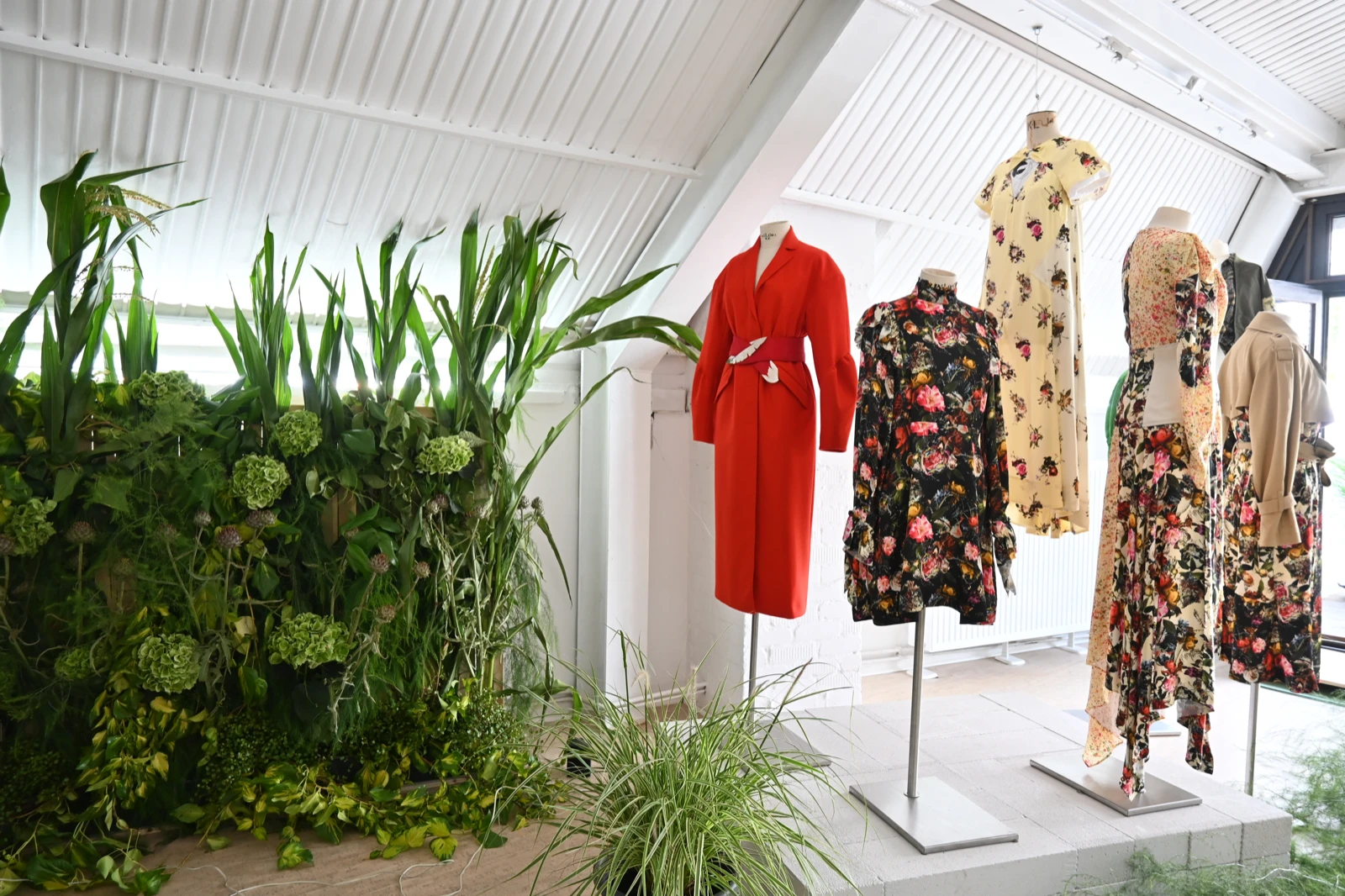 Close view of garments from Société Angelique’s collection displayed on mannequins at wildpalms: a red belted coat, a black floral jacket, a yellow floral dress, and patterned floral dresses, beside a wall of plants
