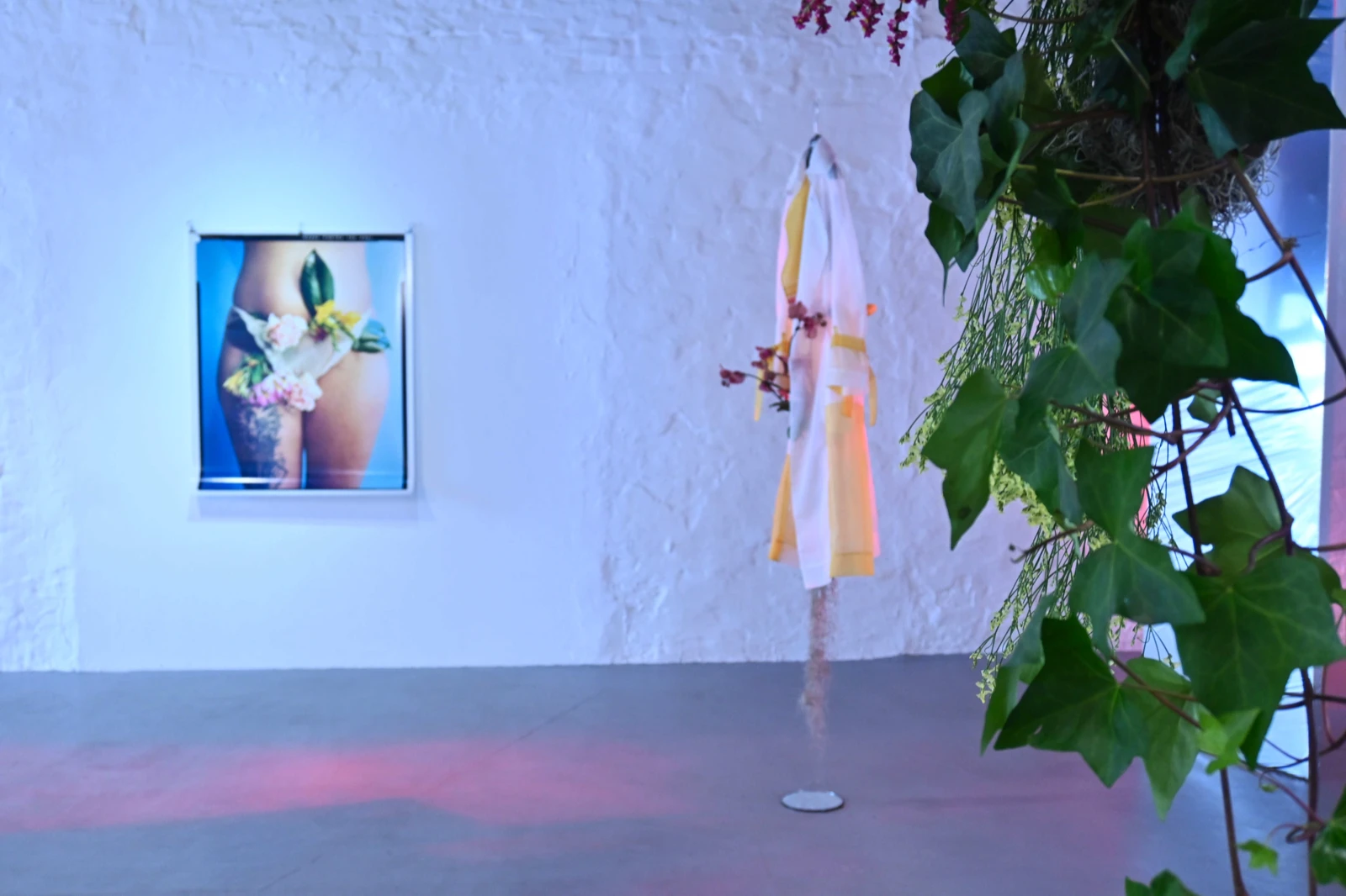 A framed photograph of a tattooed thigh adorned with flowers on a white brick wall beside a translucent white and yellow garment on a stand, with dried flowering branches and ivy nearby.