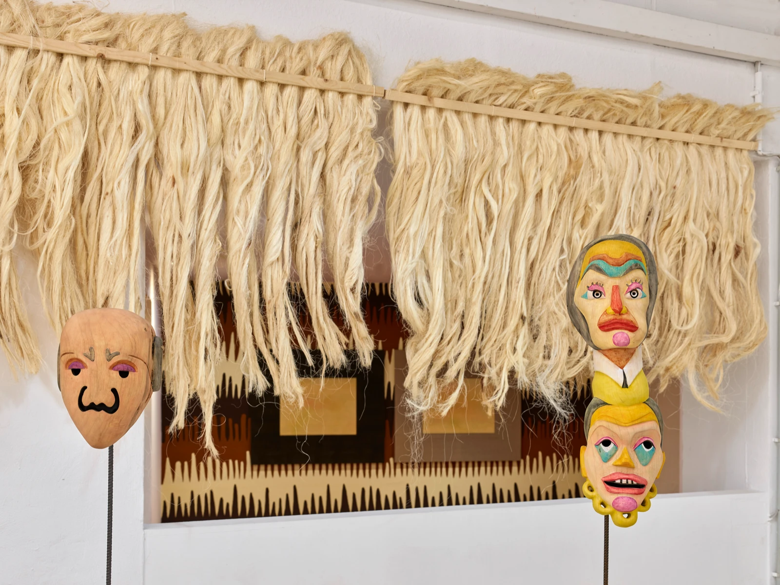 Dos máscaras de madera pintadas de Mauricio Limón bajo una cortina de fibra tejida, vista de instalación en wildpalms