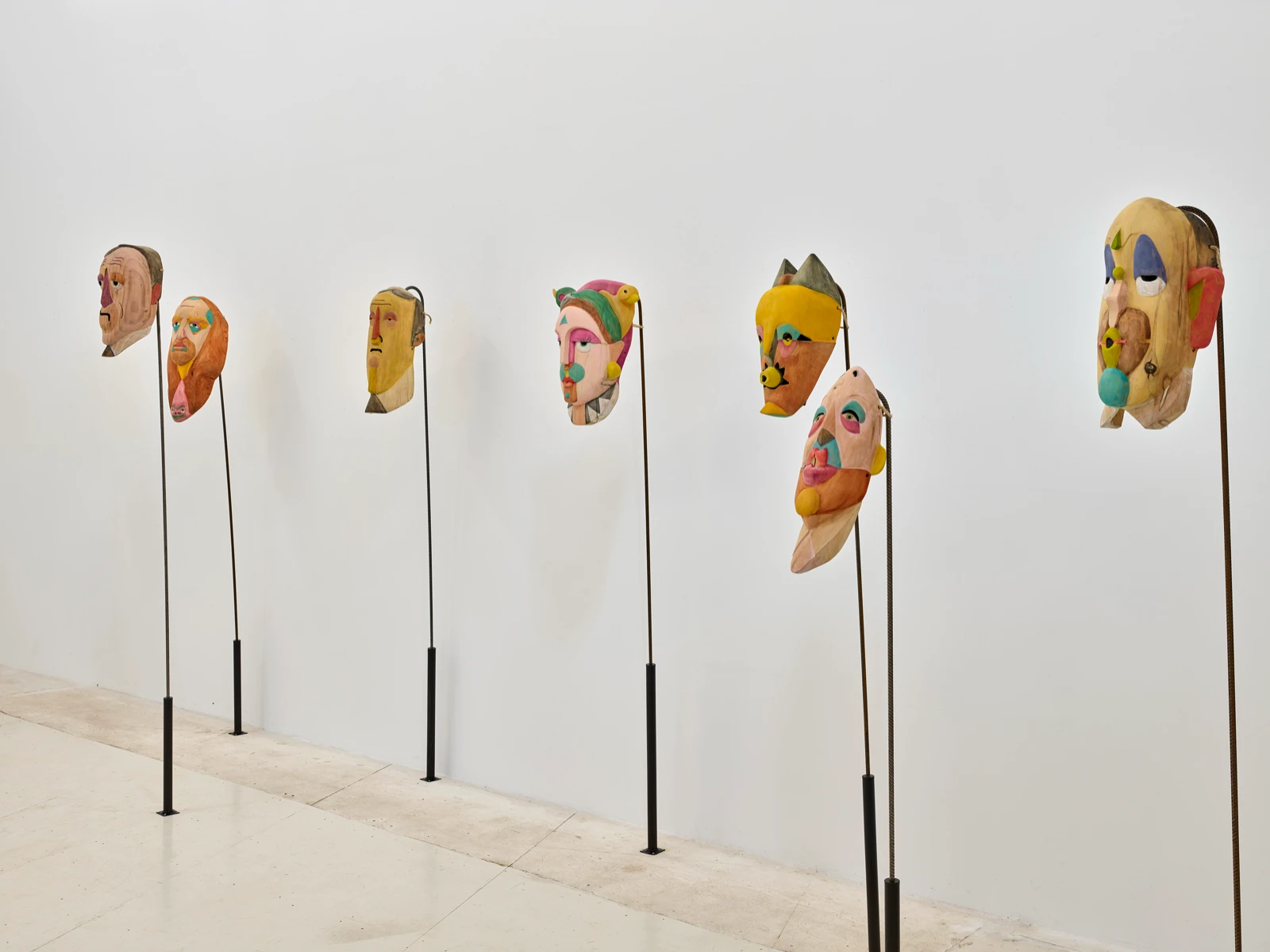 Hilera de máscaras de madera pintadas de Mauricio Limón en varillas contra una pared blanca en wildpalms