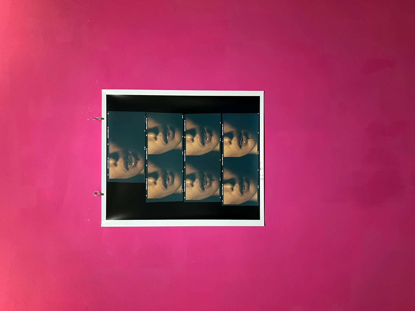A framed photographic contact sheet of repeated close-up mouth images mounted on a hot pink gallery wall.