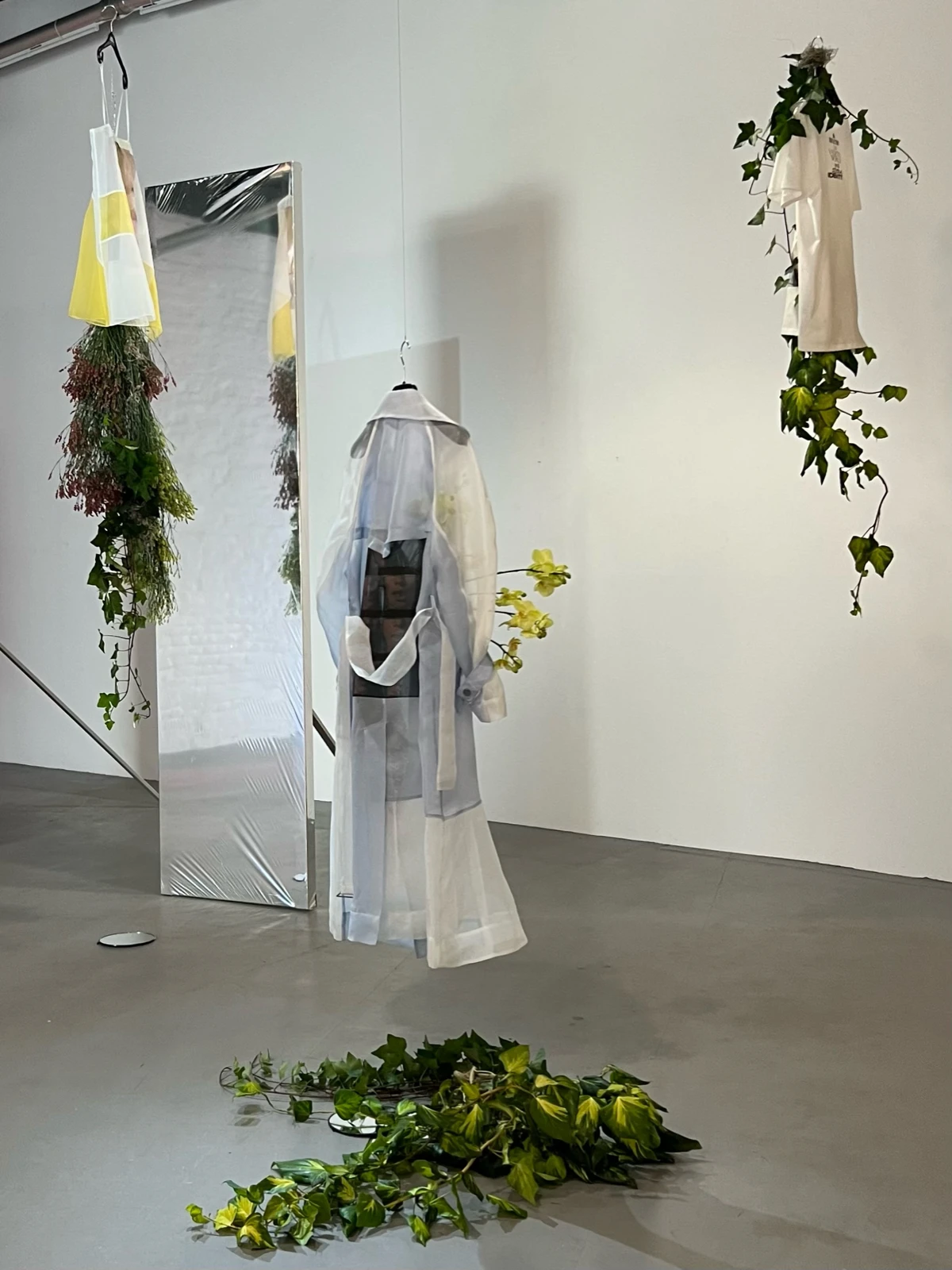 Installation view of three suspended garments: a yellow robe entwined with ivy, a sheer white robe with a printed photographic panel and ivy at its base, and a white T-shirt hanging amid trailing ivy on the wall.