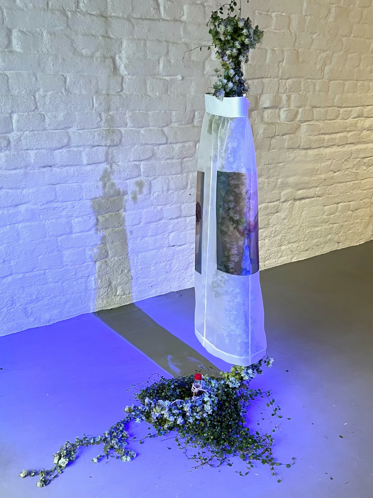 A tall white fabric sculpture printed with photographic imagery, crowned with artificial flowers and spilling into a bed of white flowers and greenery on the floor, lit in blue.