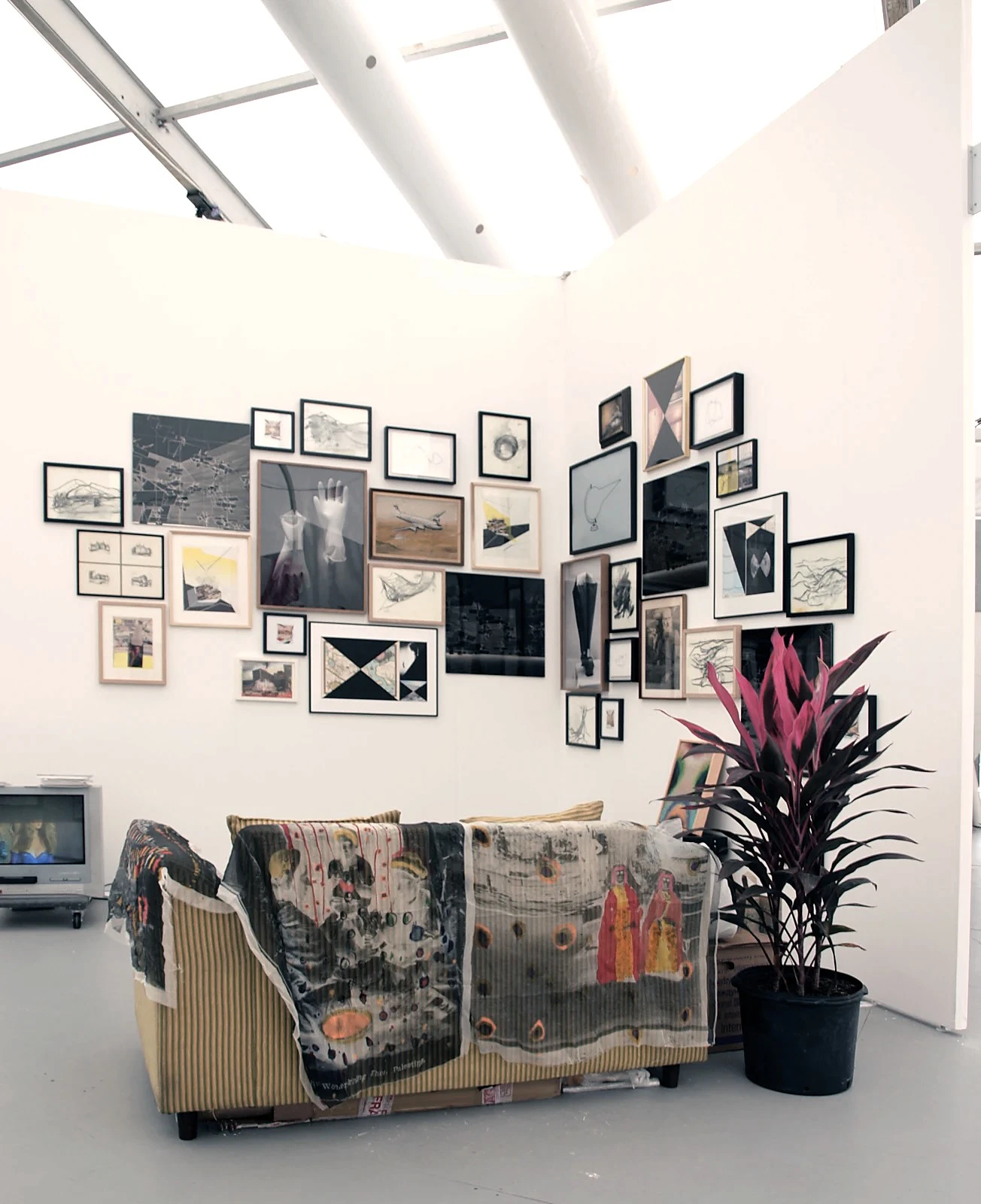 Vista de instalación de la caseta ‘La Villa’ de wildpalms en Untitled Art Fair, Miami, recreando la casa compartida por Alejandro Almanza Pereda, Anibal Catalan y Jose Antonio Elguezabal — un muro tipo salón con obras sobre un sofá cubierto y un televisor
